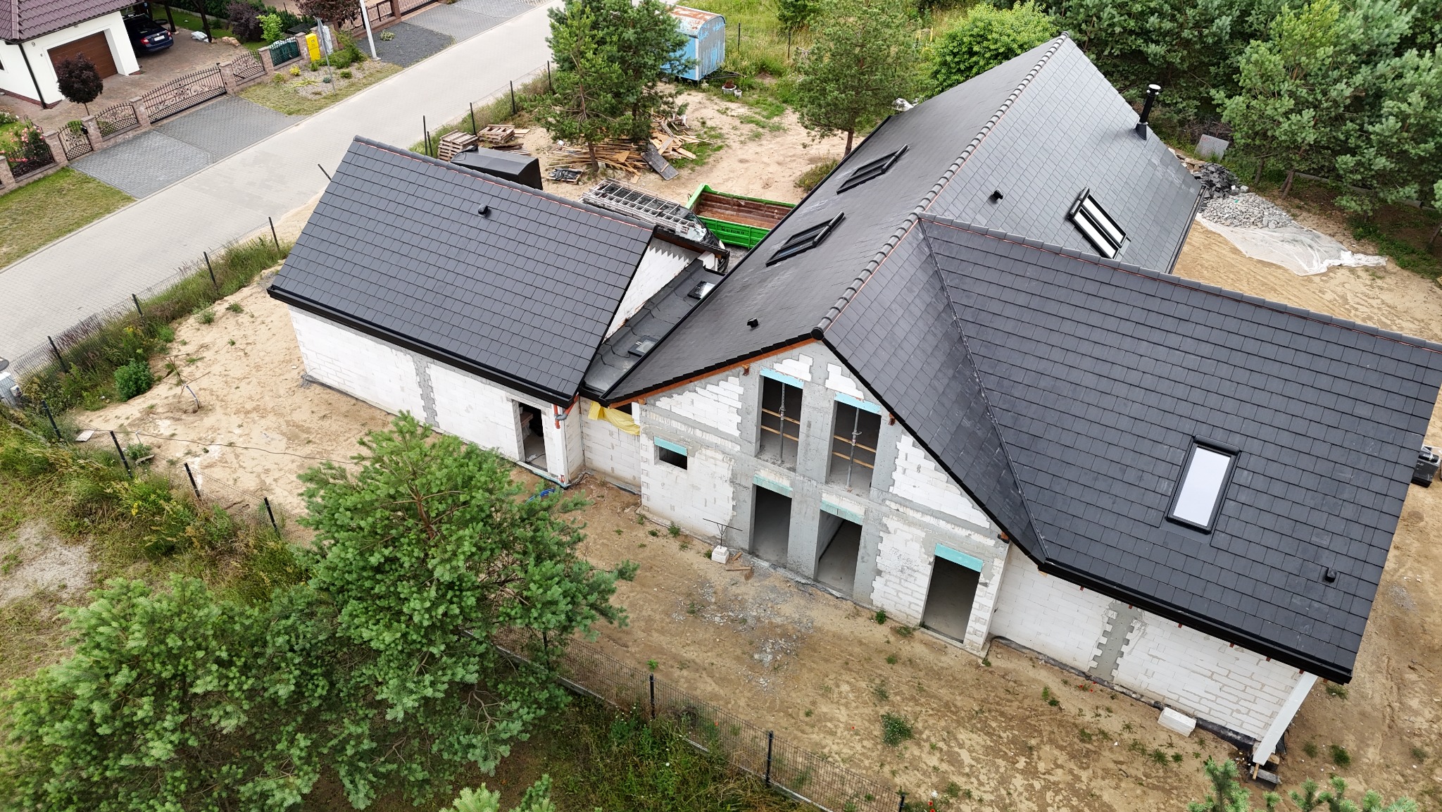 Widok z góry na budynek w trakcie budowy z częściowo ukończonym czarnym pokryciem dachowym, widoczne okna dachowe i niezabudowane otwory okienne w ścianach z betonu komórkowego, teren wokół budynku...