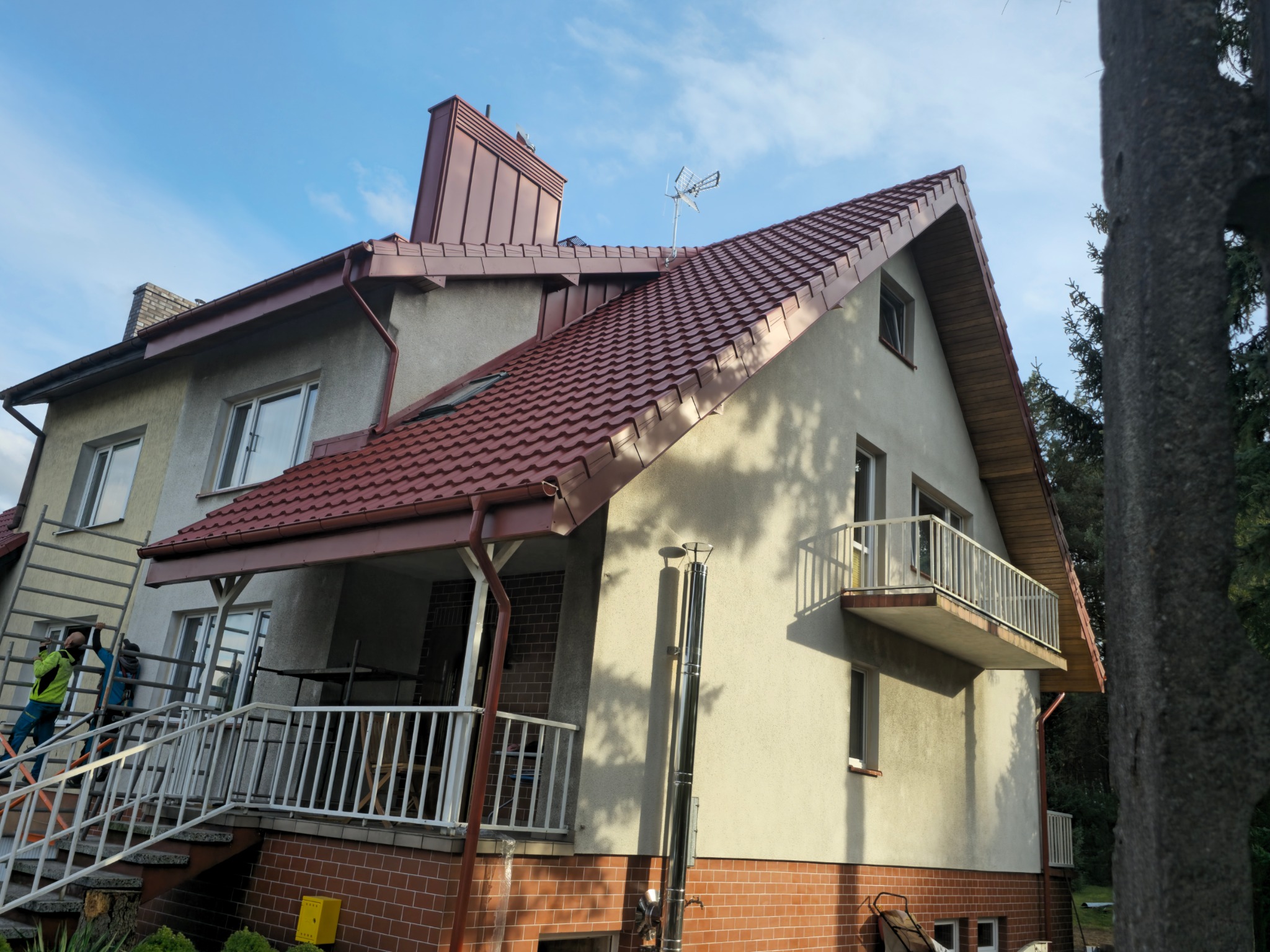 Dom jednorodzinny z nową, czerwoną dachówką i metalowym kominem, widoczni pracownicy na rusztowaniu, antena na dachu.