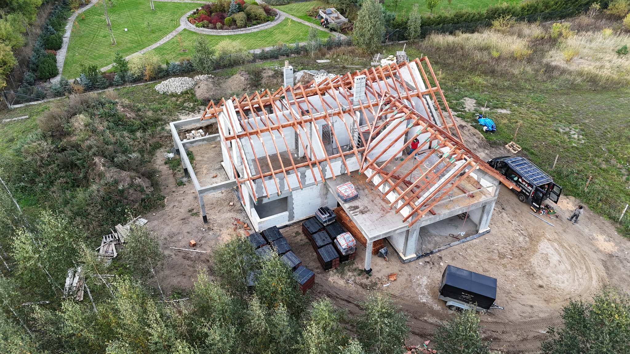 Widok z lotu ptaka na budowę domu jednorodzinnego z widoczną drewnianą konstrukcją dachu, niezabezpieczonymi otworami okiennymi, stosem dachówek na tarasie, zaparkowanym obok budowy samochodem...