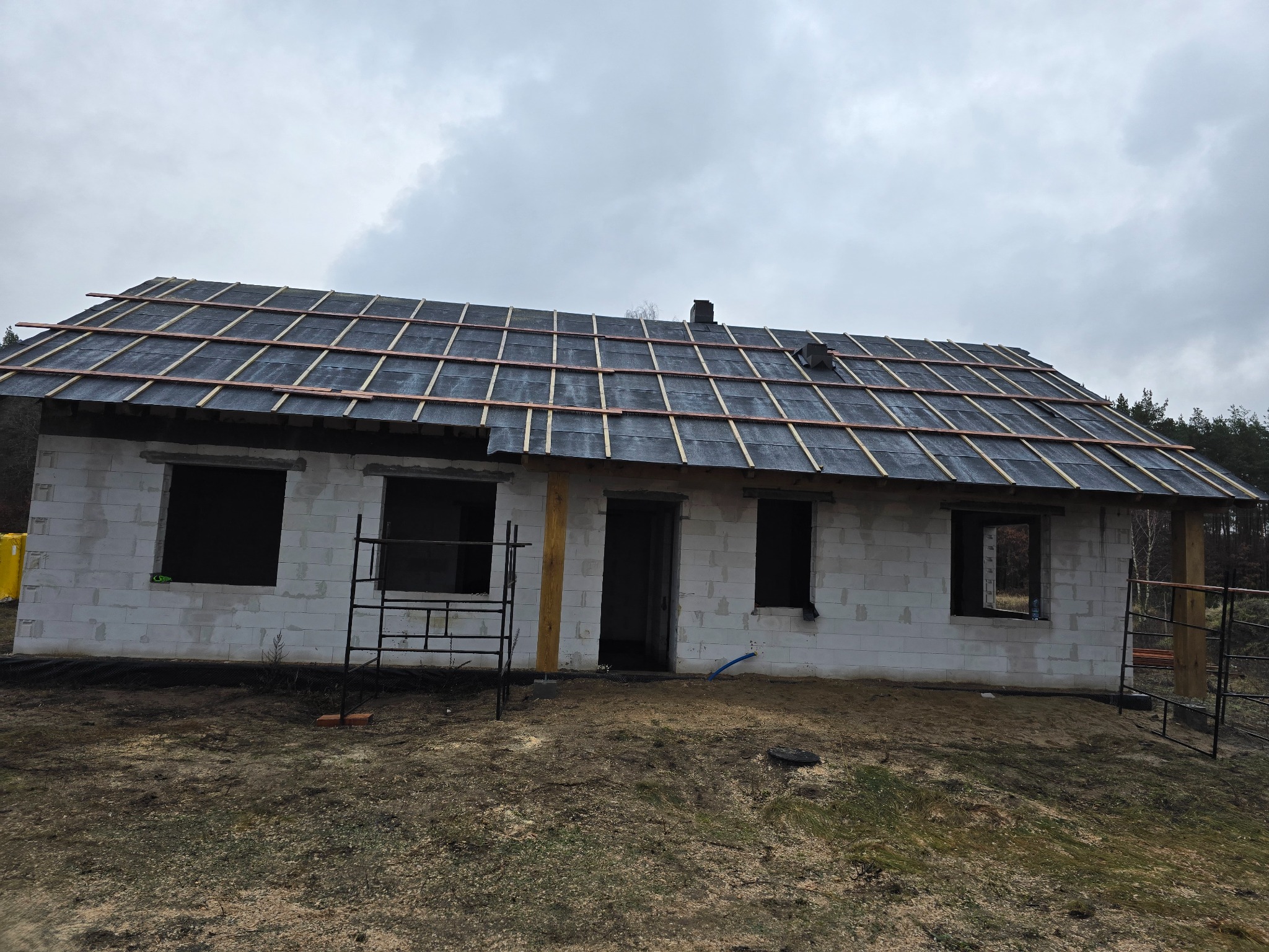 Budowa domu jednorodzinnego w trakcie: widoczna surowa konstrukcja dachu z deskowaniem i łatami, białe ściany z bloczków, czarne ramy okienne, szare niebo w tle.