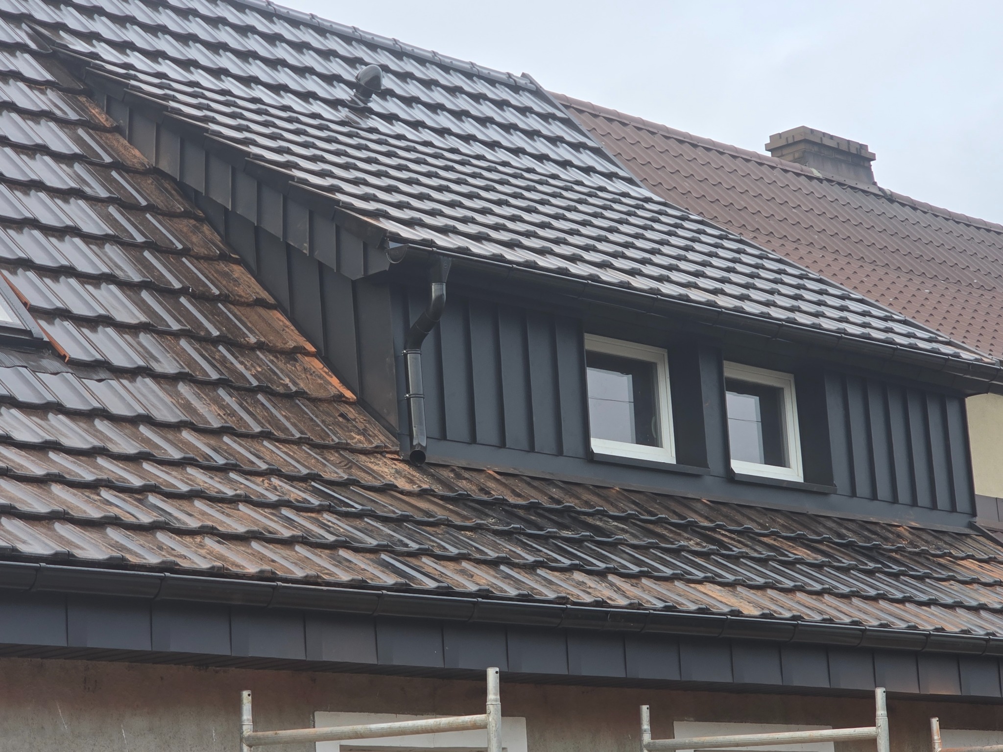 Częściowy widok dachu z ciemnej dachówki ceramicznej, z oknami dachowymi i rynną, na tle innego dachu z ceglanej dachówki. Widoczne rusztowanie.