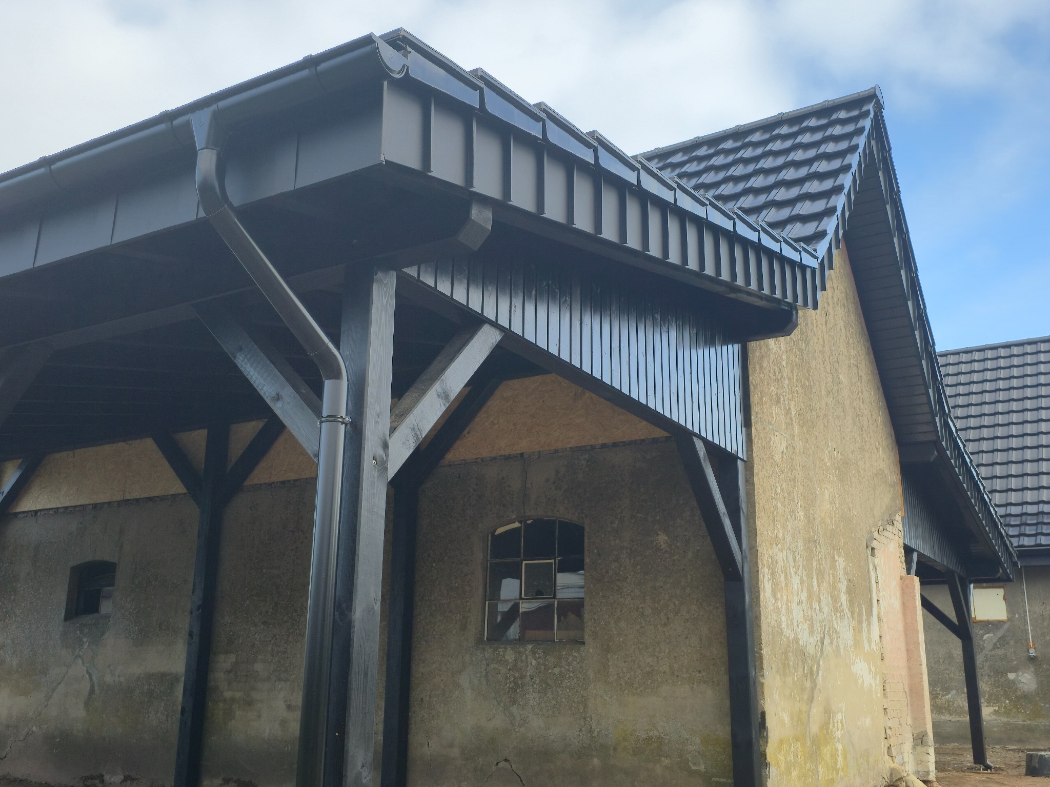 Ciemna blacha na dachu budynku z drewnianą konstrukcją, widoczne rynny i dachówka. Budynek otynkowany z małym oknem. Ujęcie z dołu.