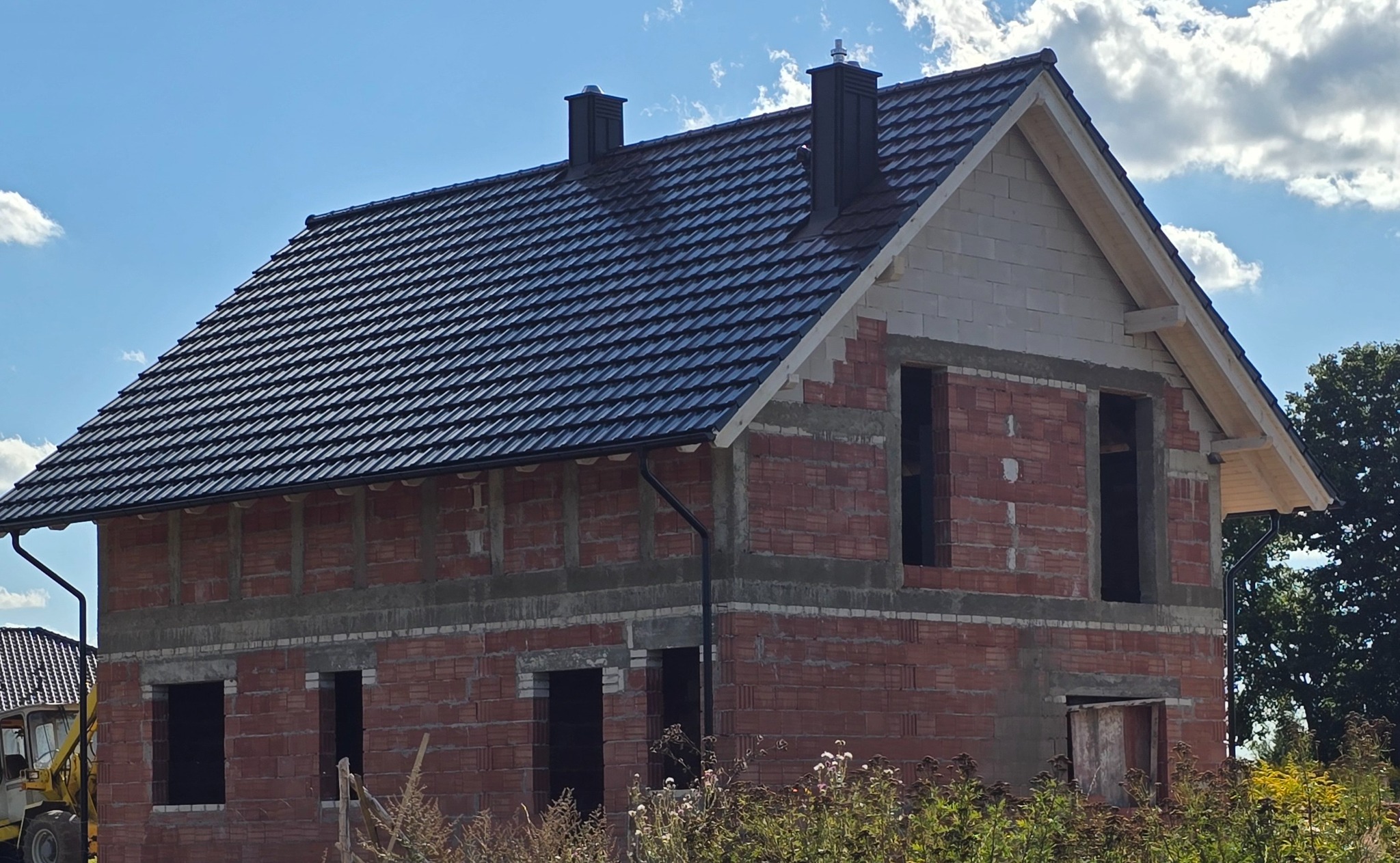 Dom w trakcie budowy z ciemnym dachem z dachówki, widoczne kominy i rynny. Budynek z czerwonej cegły, okna bez szyb. Niebo z chmurami w tle.