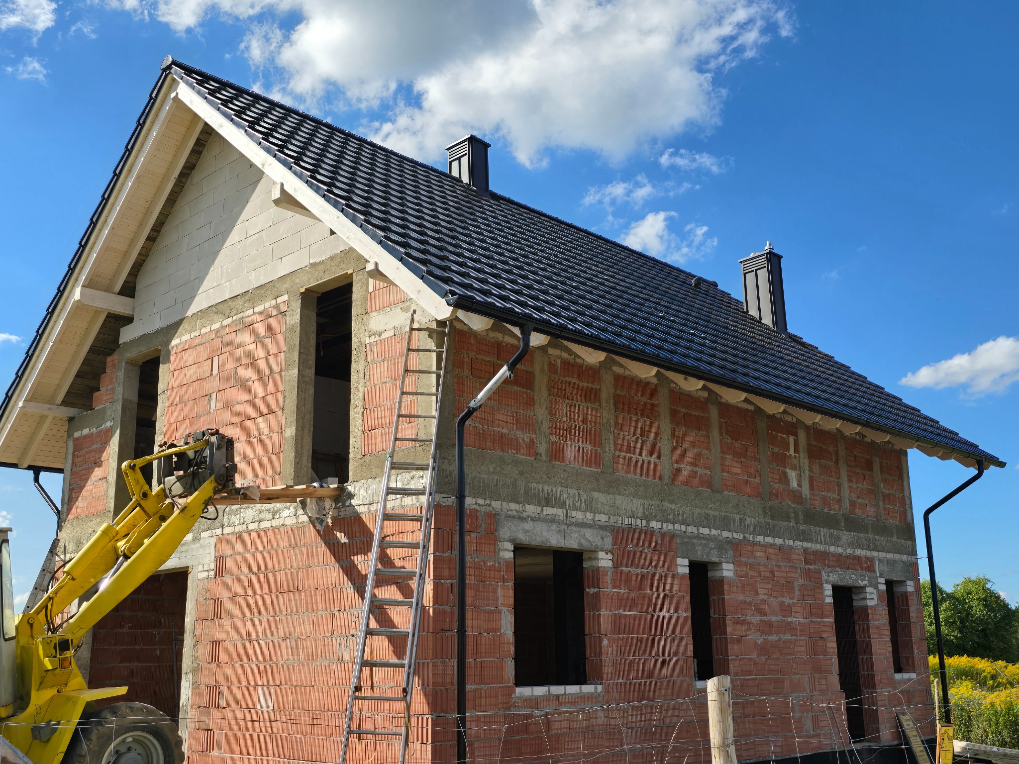 Dom w trakcie budowy z czarnym dachem i żółtą maszyną budowlaną. Widoczne cegły, okna bez szyb i drabina oparta o ścianę. Błękitne niebo w tle.