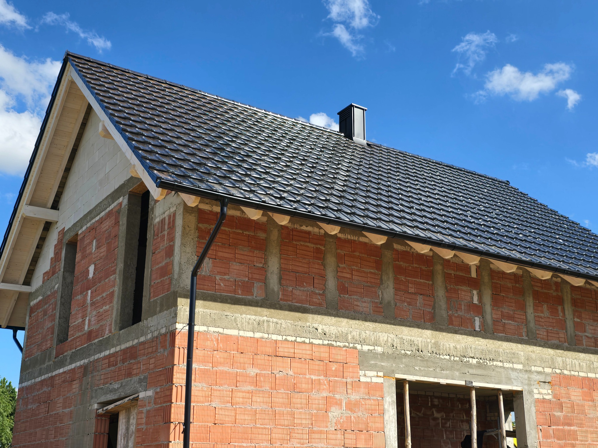 Dom w trakcie budowy z widocznym dachem pokrytym czarną dachówką i zamontowanymi rynnami. Murowana konstrukcja z czerwonej cegły i betonu pod błękitnym niebem.