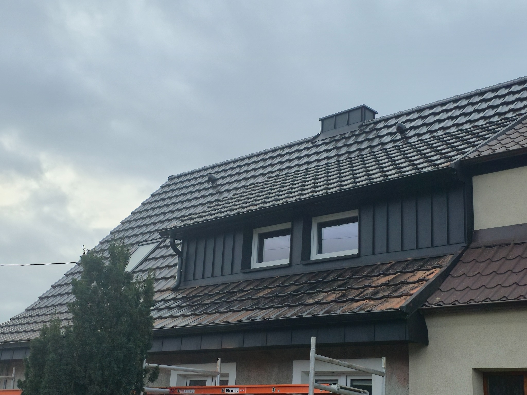 Dom z dachem pokrytym brązową dachówką ceramiczną, z nową, ciemną obróbką blacharską i orynnowaniem. Widoczne okna dachowe i fragment rusztowania.