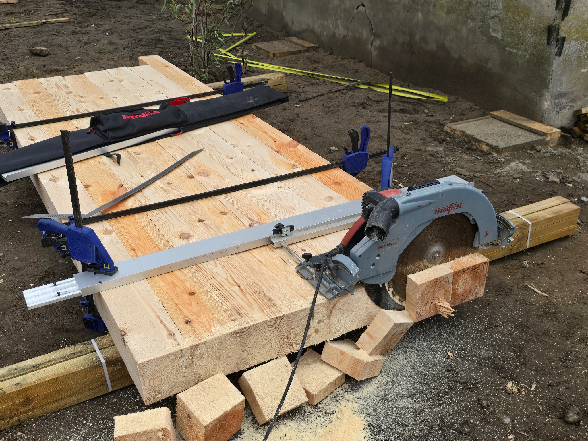Cięcie drewnianych belek piłą elektryczną Mafell, z użyciem prowadnicy i zacisków, na ziemi. Widoczne odcięte kawałki drewna i torba na narzędzia.