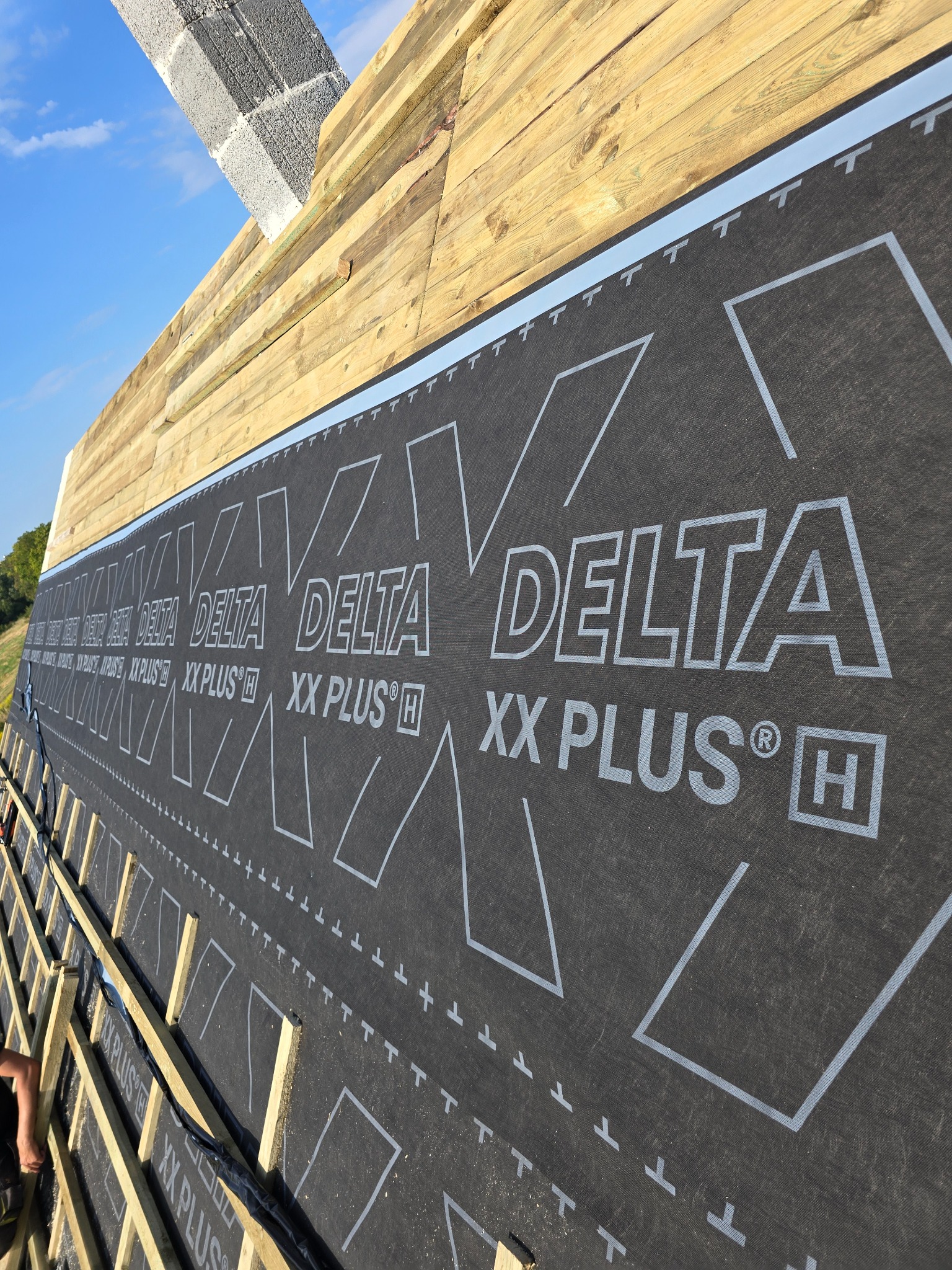 Wysoki kąt: membrana dachowa Delta XX Plus H na drewnianej więźbie dachowej, widoczne elementy konstrukcyjne i fragment nieba. Detal montażu membrany.