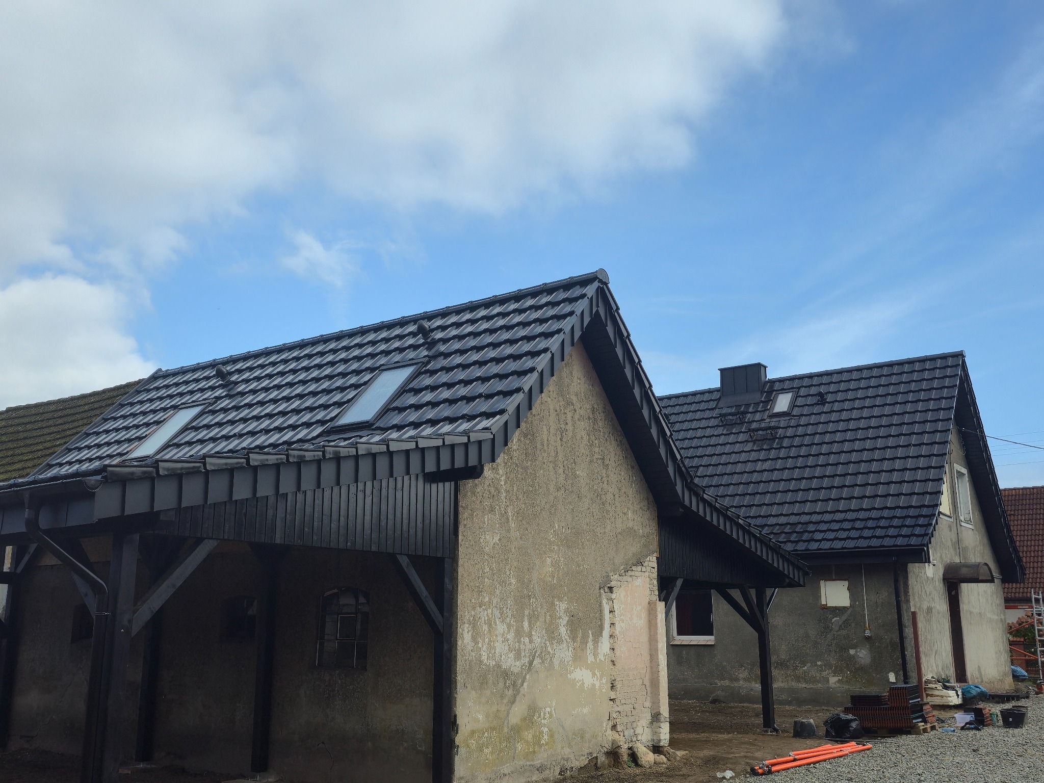 Dwa budynki z nowym, czarnym dachem z dachówką i oknami dachowymi. Jeden z budynków posiada drewnianą konstrukcję wspartą na słupach. Błękitne niebo z chmurami w tle.