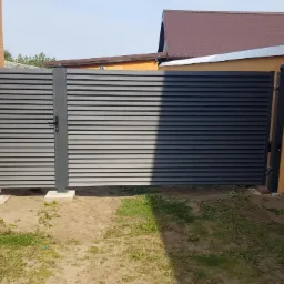 Nowoczesna, pozioma brama lamelowa w kolorze grafitowym, zamontowana na betonowych fundamentach, z widocznymi elementami mocującymi i kulistymi zakończeniami słupków.