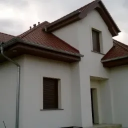 Biały dom jednorodzinny z brązowym dachem i garażem, widok z boku. Elewacja wykończona, okna z roletami zewnętrznymi, pochmurne niebo w tle.