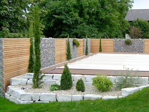 Nowoczesne ogrodzenie panelowe z drewnianymi przęsłami i gabionami, oddzielające ogród z białymi kamieniami i niskimi roślinami od trawnika.