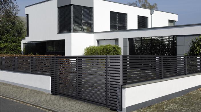 Nowoczesne ogrodzenie panelowe w kolorze antracytowym, zamontowane na niskim murku przed białym, dwukondygnacyjnym domem z płaskim dachem i żaluzjami w oknach.