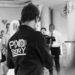 Czarno-białe ujęcie zza pleców mężczyzny z kamerą i napisem 'PIXO STORY' na kurtce, filmującego parę młodą i gości weselnych w sali.