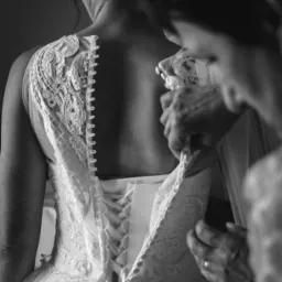 Czarno-białe zdjęcie z bliska, przedstawiające zapinanie koronkowej sukni ślubnej z wiązaniem gorsetowym na plecach panny młodej, z widocznymi dłońmi osoby pomagającej.