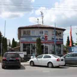 Fasada dwupiętrowego budynku firmy 'Technika Grzewcza i Sanitarna' z reklamami ceramiki, armatury, mebli i akcesoriów, z antenami na dachu i zaparkowanymi samochodami przed budynkiem.