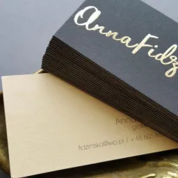 Stos wizytówek z czarnego papieru z tłoczonym złotym napisem 'Anna Fidzińska' leżący na dekoracyjnej, metalowej podstawce. Druga wizytówka w kolorze beżowym z drobnym tekstem.