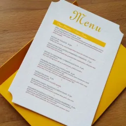 Eleganckie menu weselne z żółtym nadrukiem i ozdobnym wycięciem, częściowo wsunięte w żółtą kopertę, leżące na drewnianym stole.