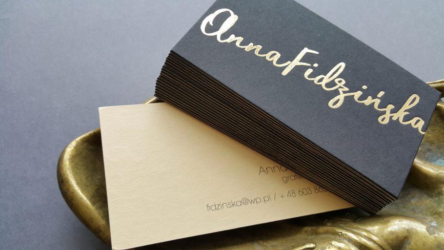 Stos wizytówek z czarnego papieru z tłoczonym złotym napisem 'Anna Fidzińska' leżący na dekoracyjnej, metalowej podstawce. Druga wizytówka w kolorze beżowym z drobnym tekstem.