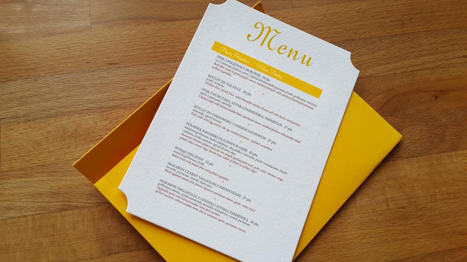 Eleganckie menu weselne z żółtym nadrukiem i ozdobnym wycięciem, częściowo wsunięte w żółtą kopertę, leżące na drewnianym stole.