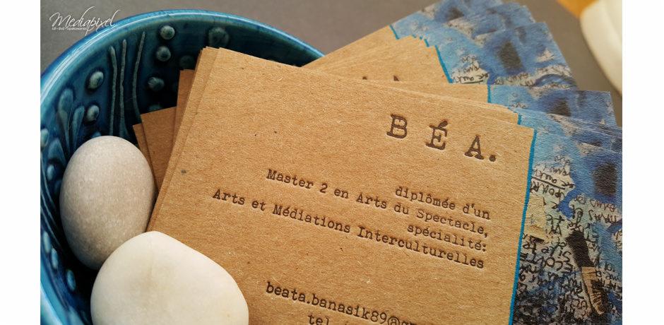 Stos wizytówek z brązowego papieru z wytłoczonym tekstem, ułożonych w błękitnej ceramicznej miseczce obok dwóch białych kamieni.