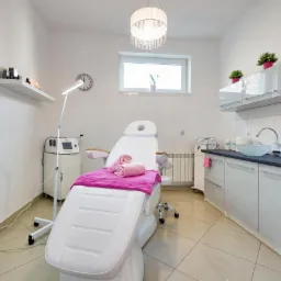 Jasne wnętrze gabinetu kosmetycznego z białym fotelem zabiegowym przykrytym różowym ręcznikiem i zwiniętymi ręcznikami, widoczny sprzęt kosmetyczny, umywalka i szafki.