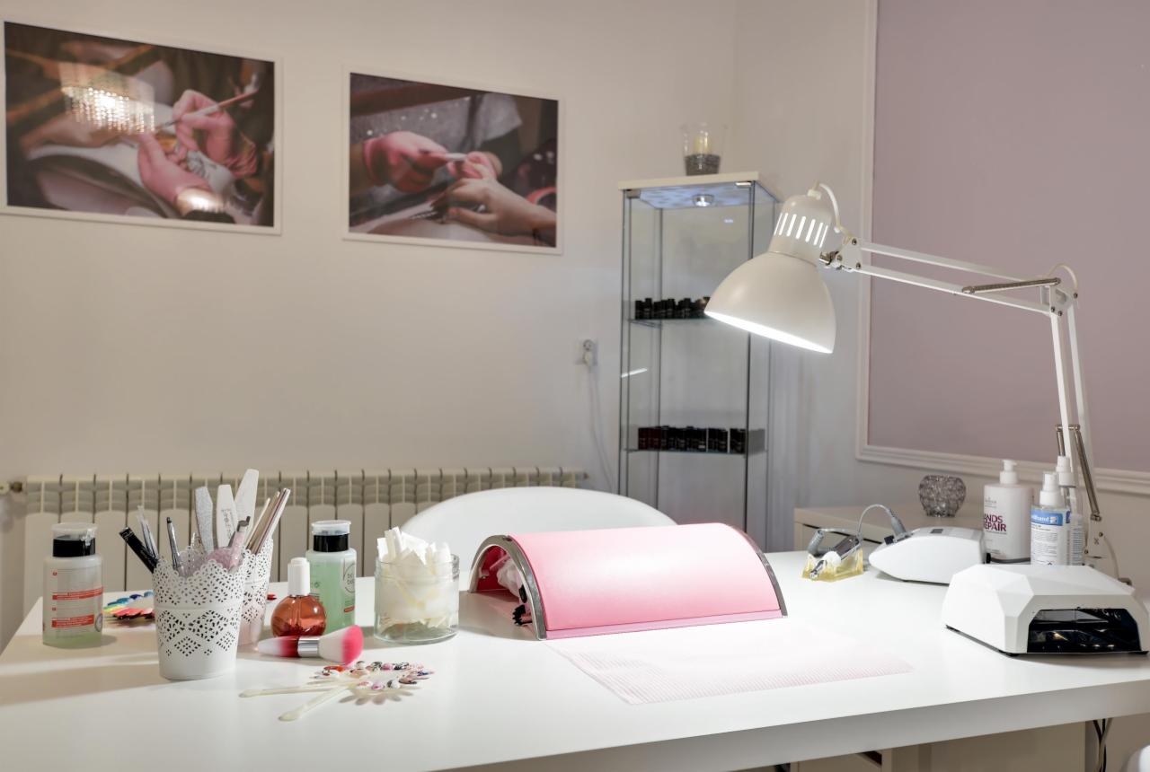 Stanowisko pracy kosmetyczki: biały blat z lampą UV do paznokci, pojemnikami z pędzlami i pilnikami, preparatami, w tle zdjęcia z manicure na ścianie i witryna z lakierami.