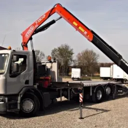 Ciężarówka Renault z hydraulicznym dźwigiem Fassi F230XP, wysięgnik podniesiony, stabilizatory opuszczone, tło stanowią białe przyczepy i budynek gospodarczy.