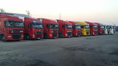 Rząd czerwonych, żółtych i białych ciężarówek zaparkowanych na szarym placu, z widocznym budynkiem w tle i drzewem bez liści po lewej stronie.