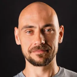 Portret łysiejącego mężczyzny z brodą, patrzącego wprost, na czarnym tle.