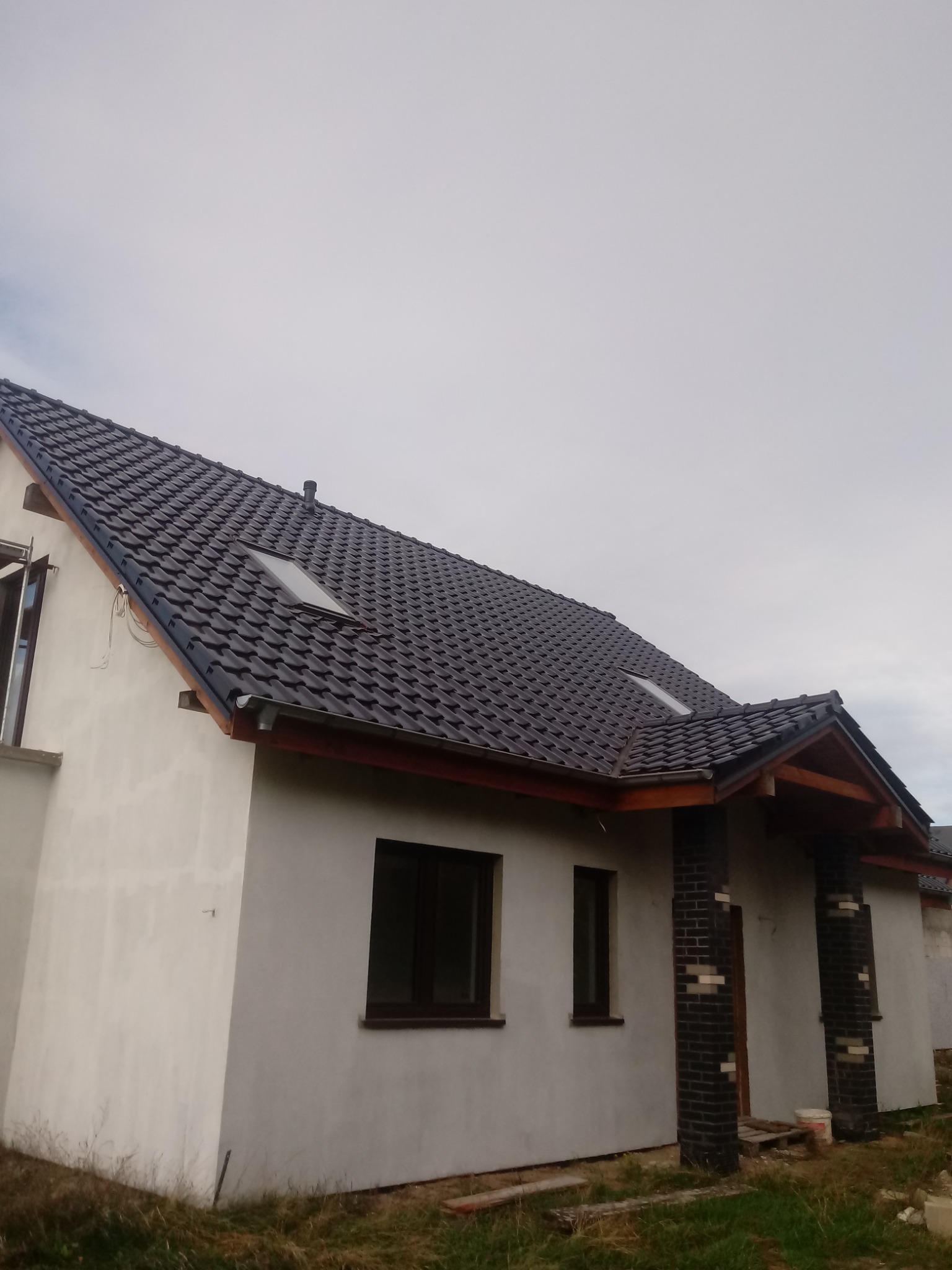 Nowy dom jednorodzinny z ciemną dachówką ceramiczną i oknami dachowymi, widok z boku na elewację w trakcie wykończenia.