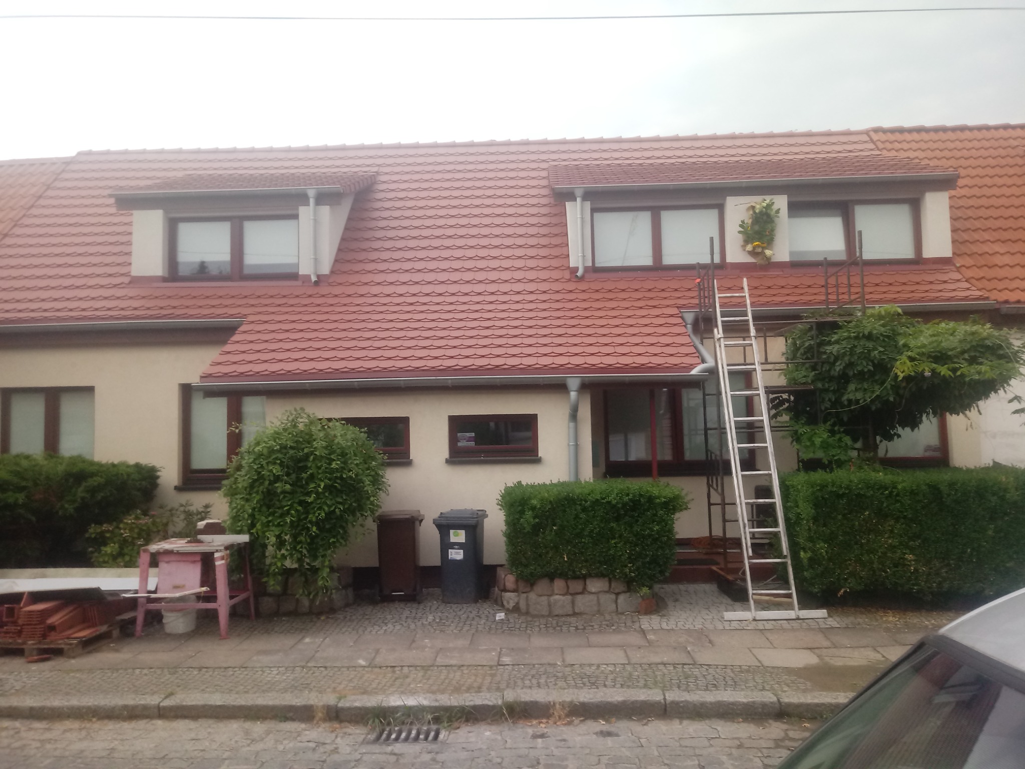 Dom z czerwoną dachówką po renowacji, z widoczną drabiną i elementami dekoracyjnymi na fasadzie, oraz stertą dachówek obok prowizorycznego stołu