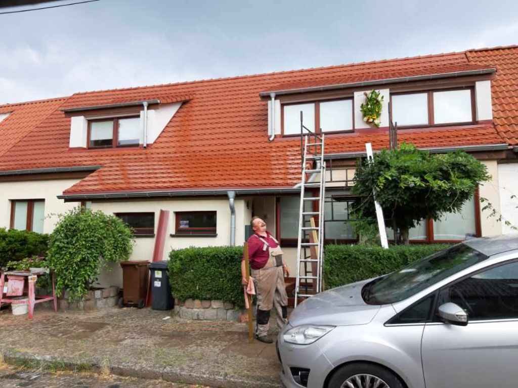 Pracownik w beżowym kombinezonie i czerwonej koszuli stoi oparty o drabinę na tle domu z czerwoną dachówką i nowymi oknami, prawdopodobnie po montażu rynien.