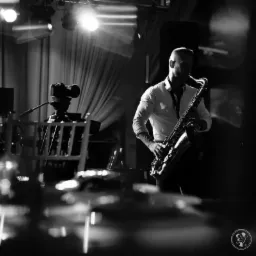 Czarno-białe zdjęcie saksofonisty grającego na scenie, z widocznym sprzętem nagłośnieniowym i oświetleniem, uchwycone z niskiego kąta.