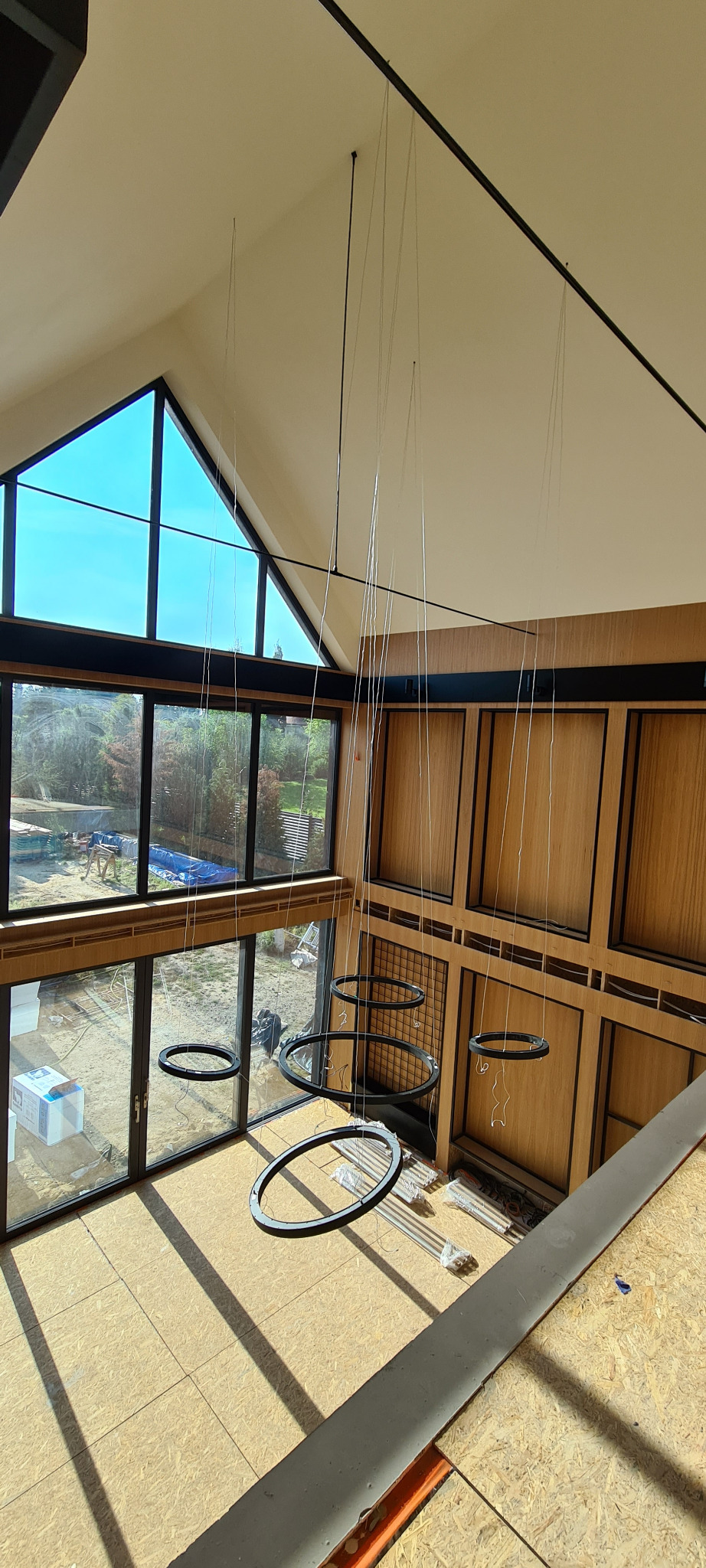 Wysoki pokój z dużymi oknami i drewnianą boazerią w trakcie remontu, widoczne wiszące okrągłe lampy bez kloszy i materiały budowlane na podłodze.