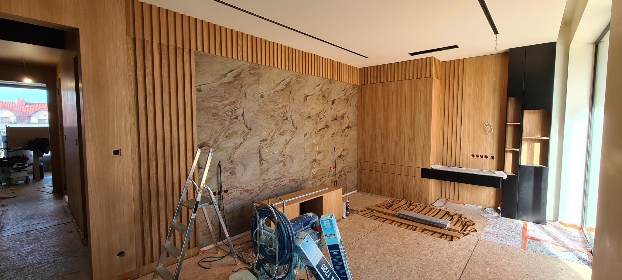 Pokój w trakcie wykańczania z panelami ściennymi z jasnego drewna, marmurową ścianą akcentującą, drabina, sprzęt i materiały budowlane na podłodze pokrytej płytami OSB.