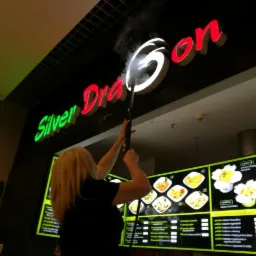 Kobieta czyści parą podświetlany szyld 'Silver Dragon' w lokalu gastronomicznym, widoczny fragment menu za nią.