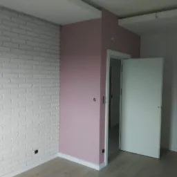 Pokój po remoncie: ściana z białej cegły dekoracyjnej kontrastuje z różową ścianą obok otwartych białych drzwi, widoczna podłoga z paneli.