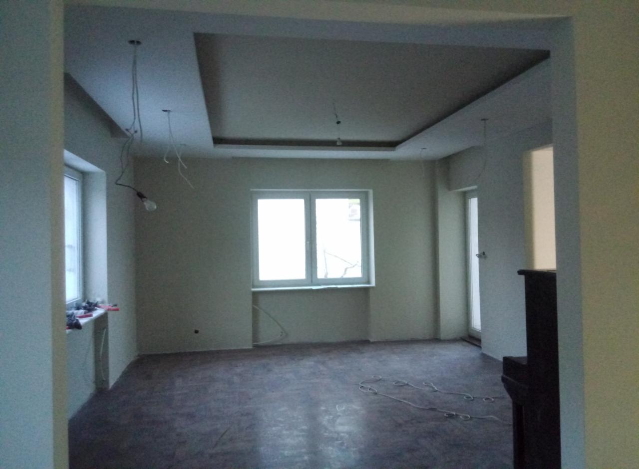 Wnętrze pokoju w trakcie prac wykończeniowych, widoczny sufit podwieszany z wystającymi kablami oświetleniowymi, przygotowane ściany do malowania, oraz częściowo ułożona podłoga