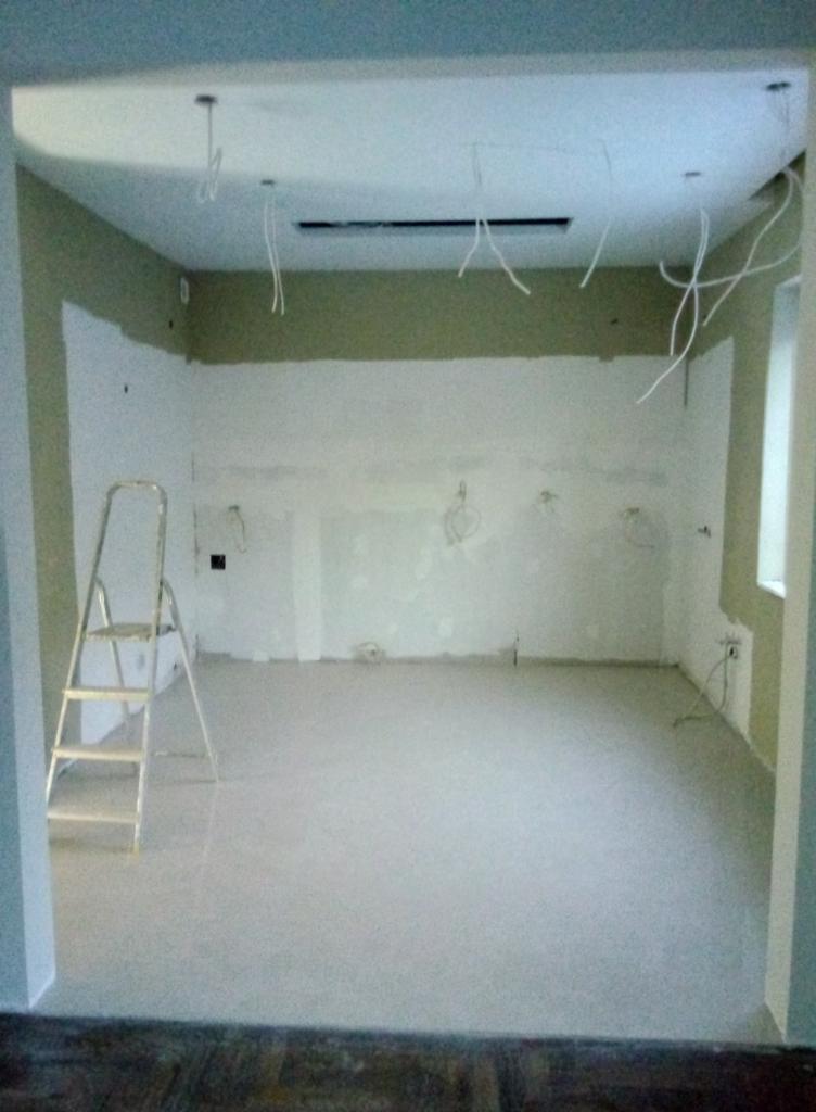 Pokój w trakcie prac wykończeniowych: ściany przygotowane pod malowanie, częściowo zagruntowane, wystające przewody elektryczne z sufitu, drabina aluminiowa przy ścianie, w tle widoczne drewniane...