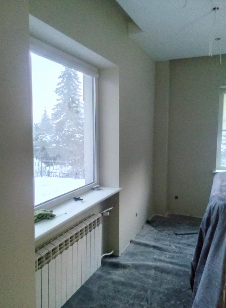 Świeżo pomalowane ściany w pokoju z oknem, zabezpieczona podłoga folią malarską, widoczny grzejnik i fragment zasłoniętego mebla.