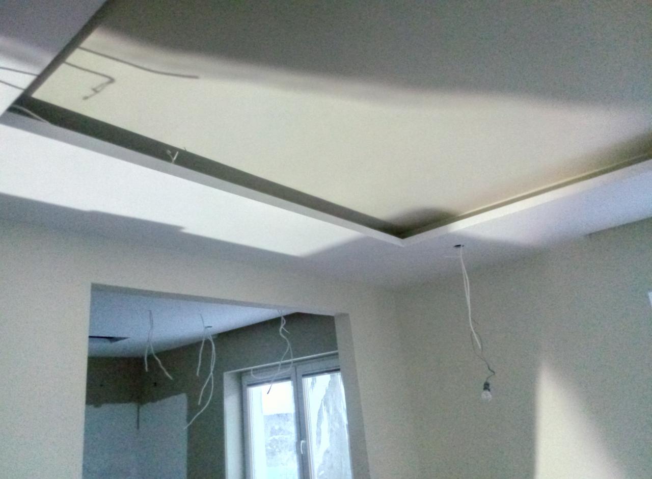 Wykończony sufit podwieszany z oświetleniem w trakcie instalacji, widoczne kable zwisające z otworu, ściany w jasnym odcieniu szarości, w tle widoczny otwór drzwiowy do innego pomieszczenia z oknem.