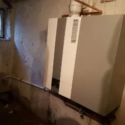 Szary piec grzewczy marki Wolf zamontowany na ścianie w piwnicy z widocznymi rurami miedzianymi i plastikowymi.