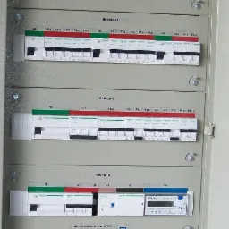 Otwarta skrzynka rozdzielcza z bezpiecznikami, wyłącznikami i licznikiem energii elektrycznej, podzielona na sekcje A-E, z żółto-zielonym przewodem ochronnym po prawej stronie.