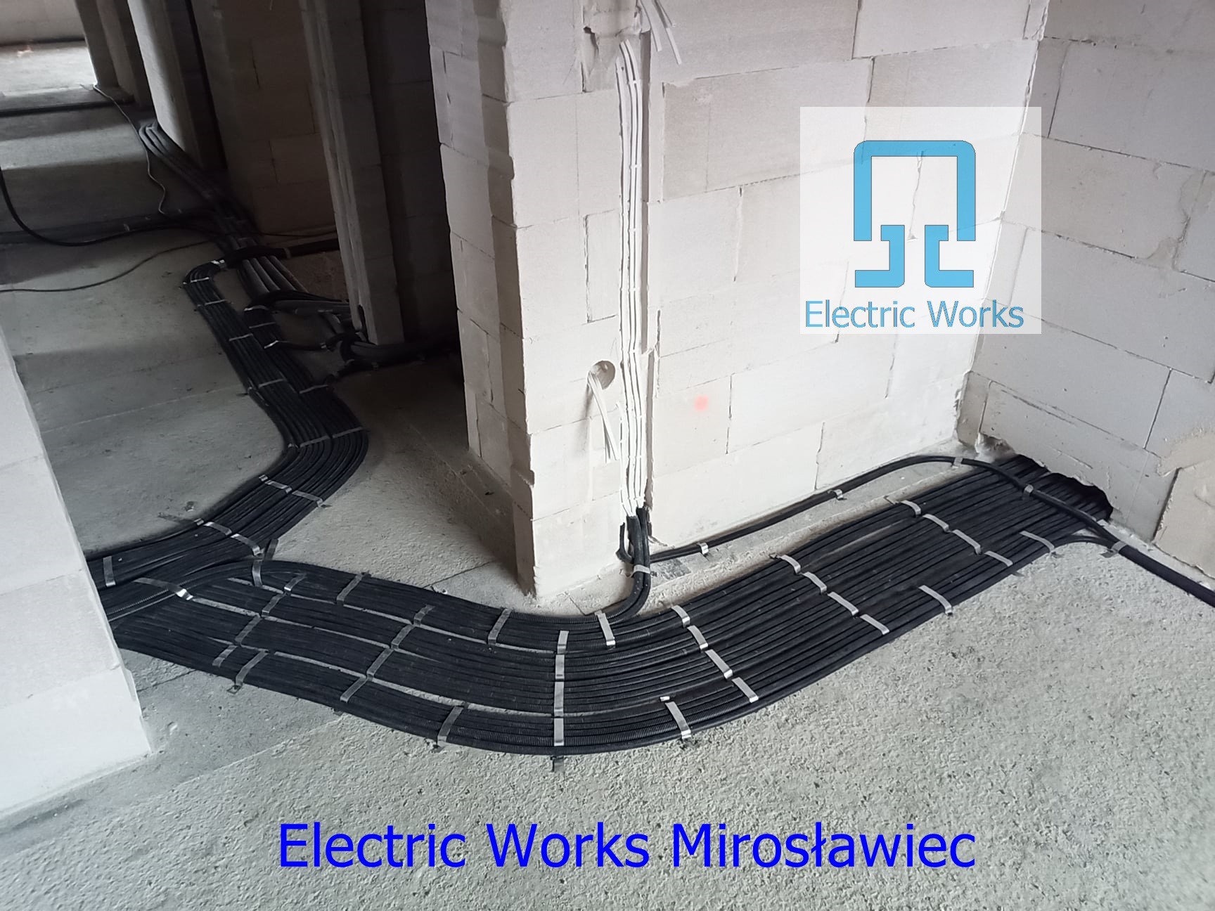 Równolegle ułożone rury instalacyjne w peszlu, przymocowane metalowymi uchwytami do surowego betonowego podłoża w budynku w trakcie budowy, z widocznymi fragmentami ścian z białego betonu...