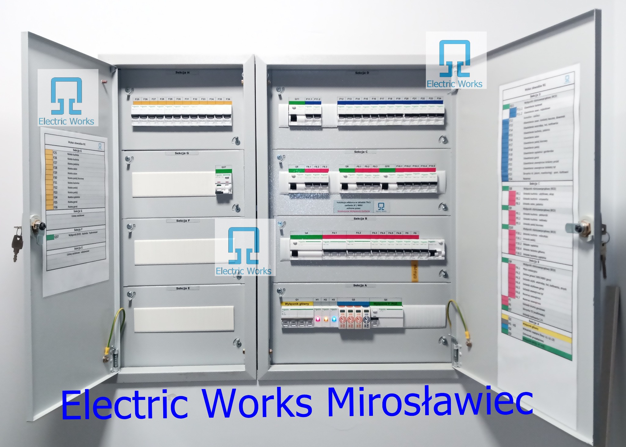 Otwarta szafa rozdzielcza z bezpiecznikami, opisana sekcjami A-H, z listą obwodów na drzwiach, widoczne zabezpieczenia i wyłączniki, klucze w zamku.