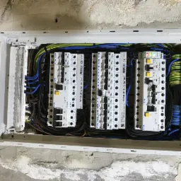 Otwarta skrzynka z bezpiecznikami i przewodami w trakcie montażu w surowej ścianie, widoczne liczne kable w różnych kolorach i białe moduły zabezpieczeń firmy Eaton.