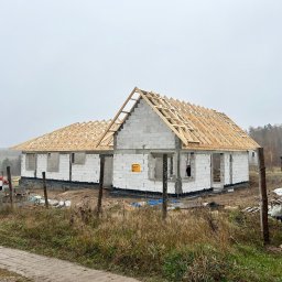 RJ-Dach - Surowy stan domu jednorodzinnego z białych bloczków, z widoczną drewnianą konstrukcją dachu w trakcie budowy, w tle krajobraz polny i pochmurne niebo.