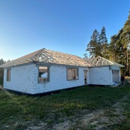 RJ-Dach - Surowy stan domu z białych bloczków, z częściowo ukończoną drewnianą konstrukcją dachu, w słoneczny dzień, na tle zieleni.