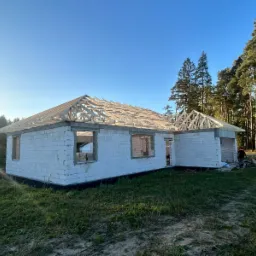 Surowy stan domu z białych bloczków, z częściowo ukończoną drewnianą konstrukcją dachu, w słoneczny dzień, na tle zieleni.